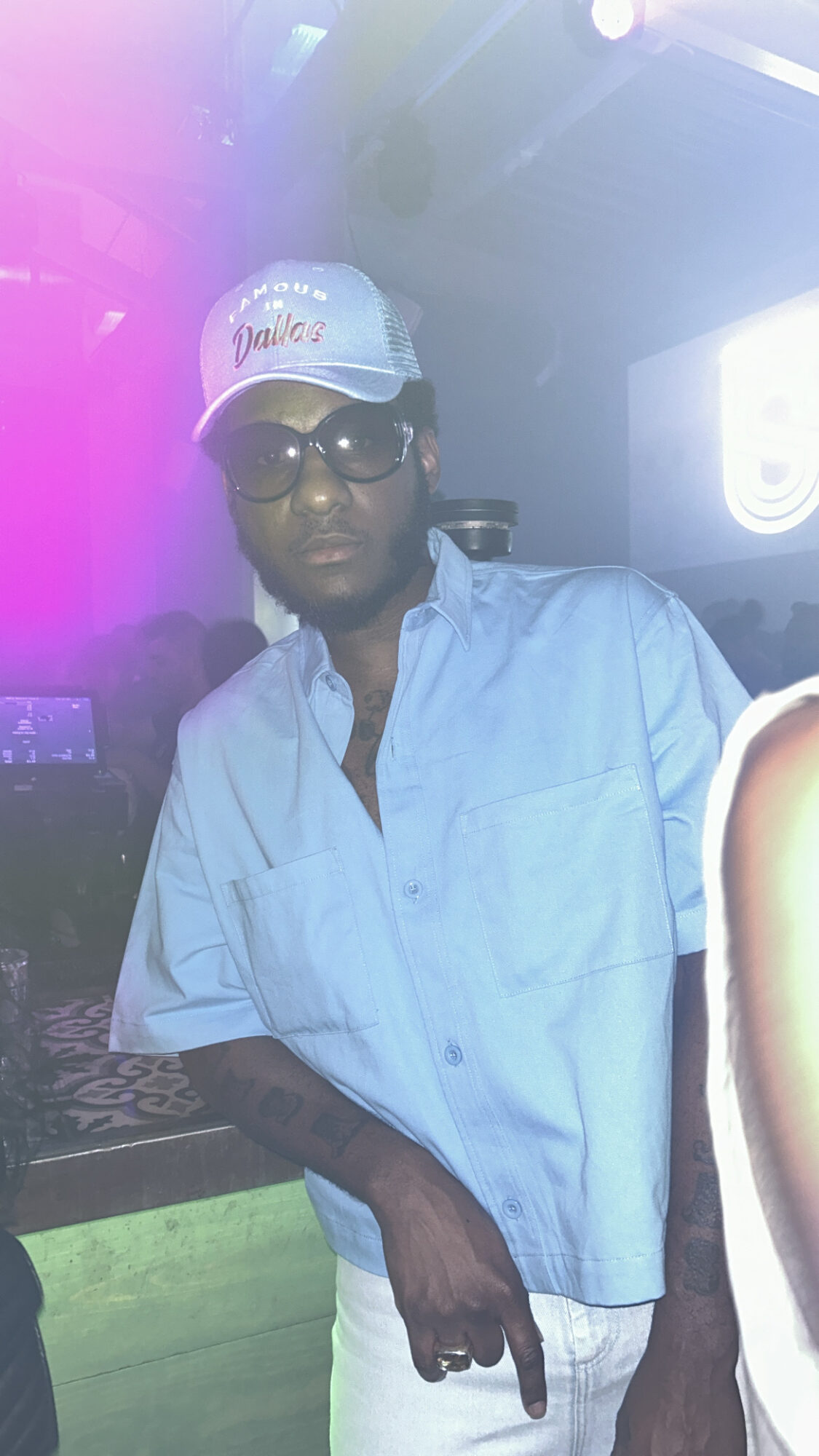 Person wearing glasses, a cap, and a light blue shirt in a dimly lit environment with colorful lights.