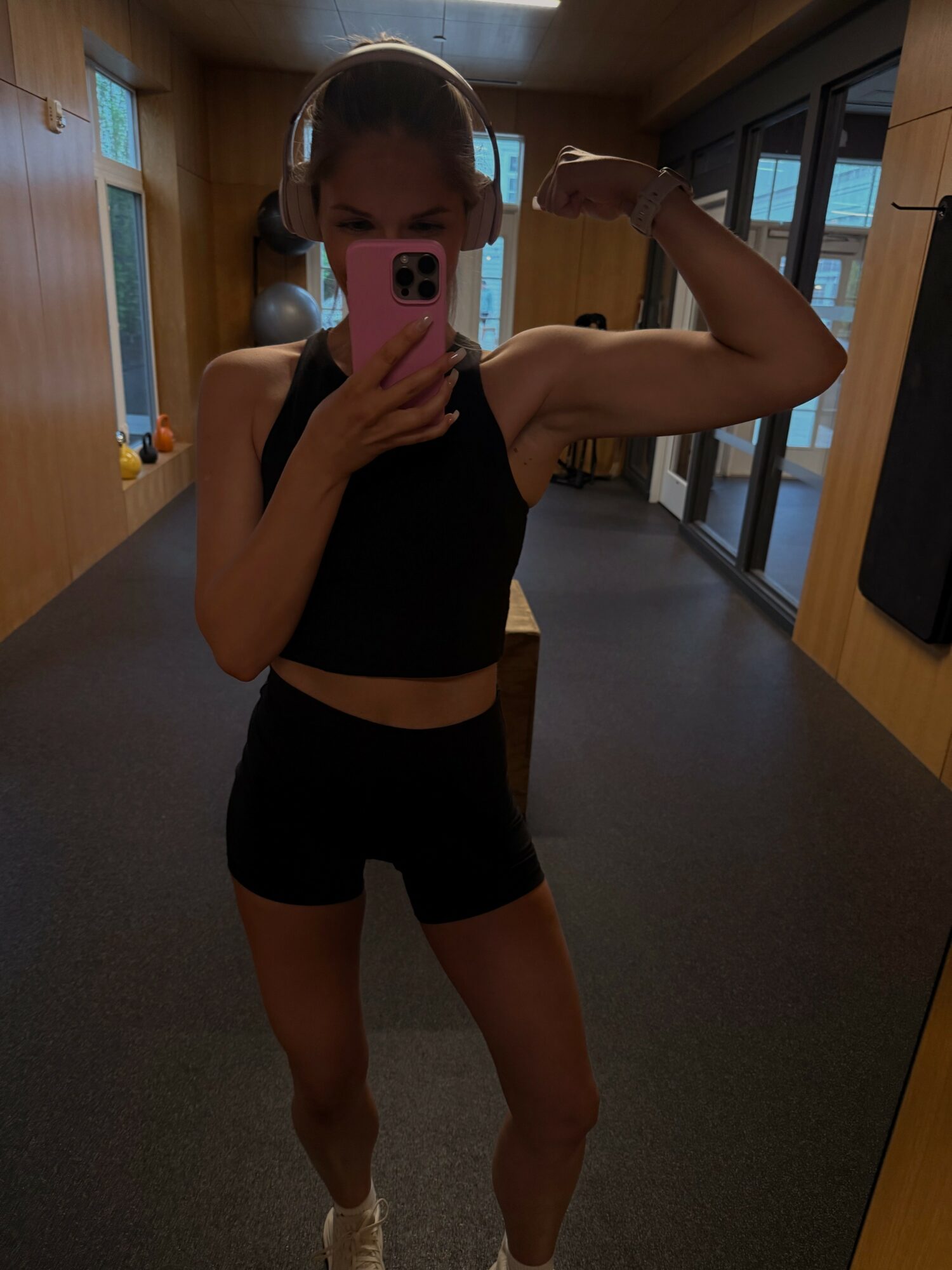 Person in gym mirror taking a selfie, flexing arm, wearing headphones, black sports bra, and shorts.
