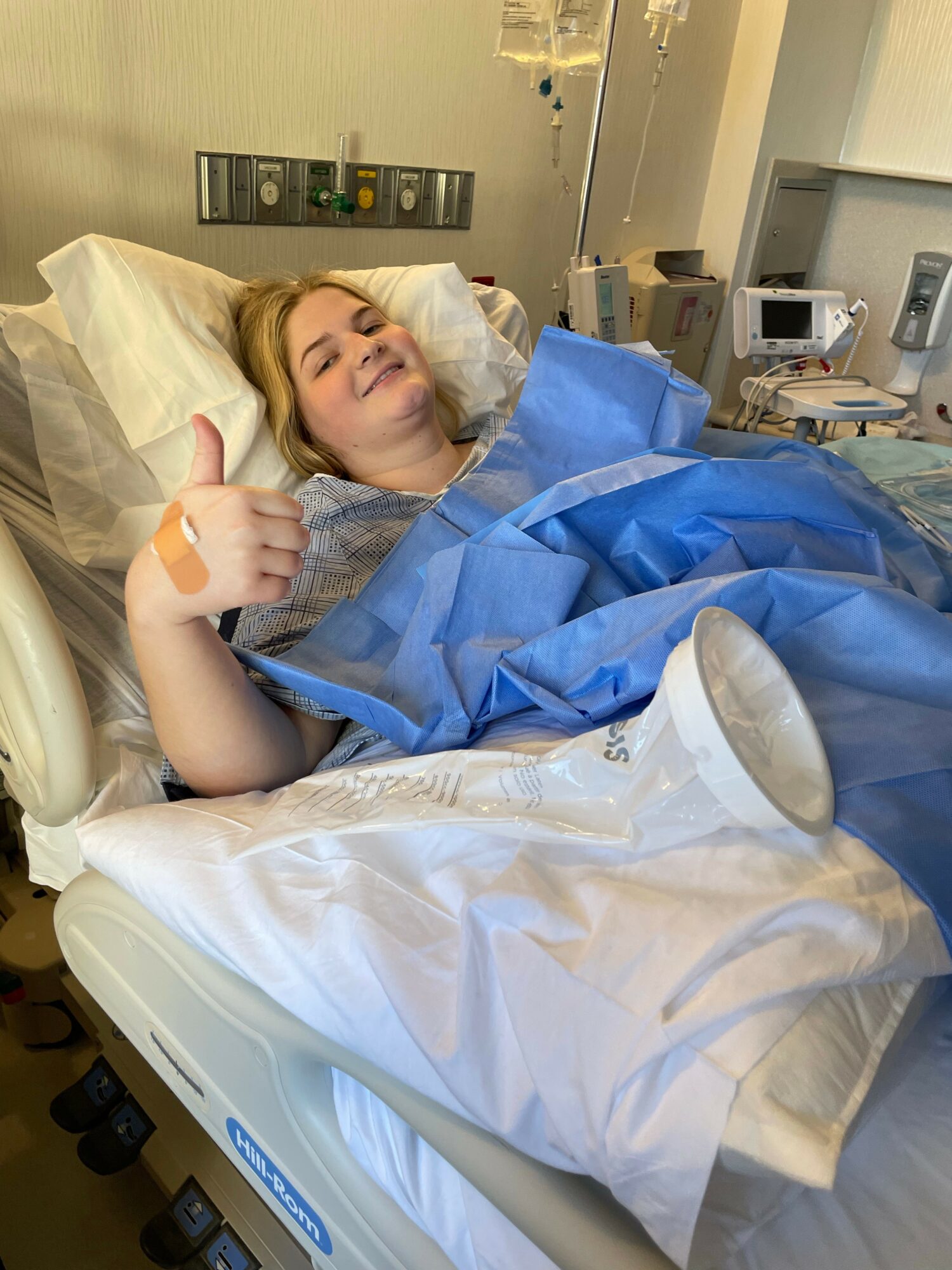 Woman lying in hospital bed giving a thumbs-up, covered with blue medical sheets, in a hospital room.