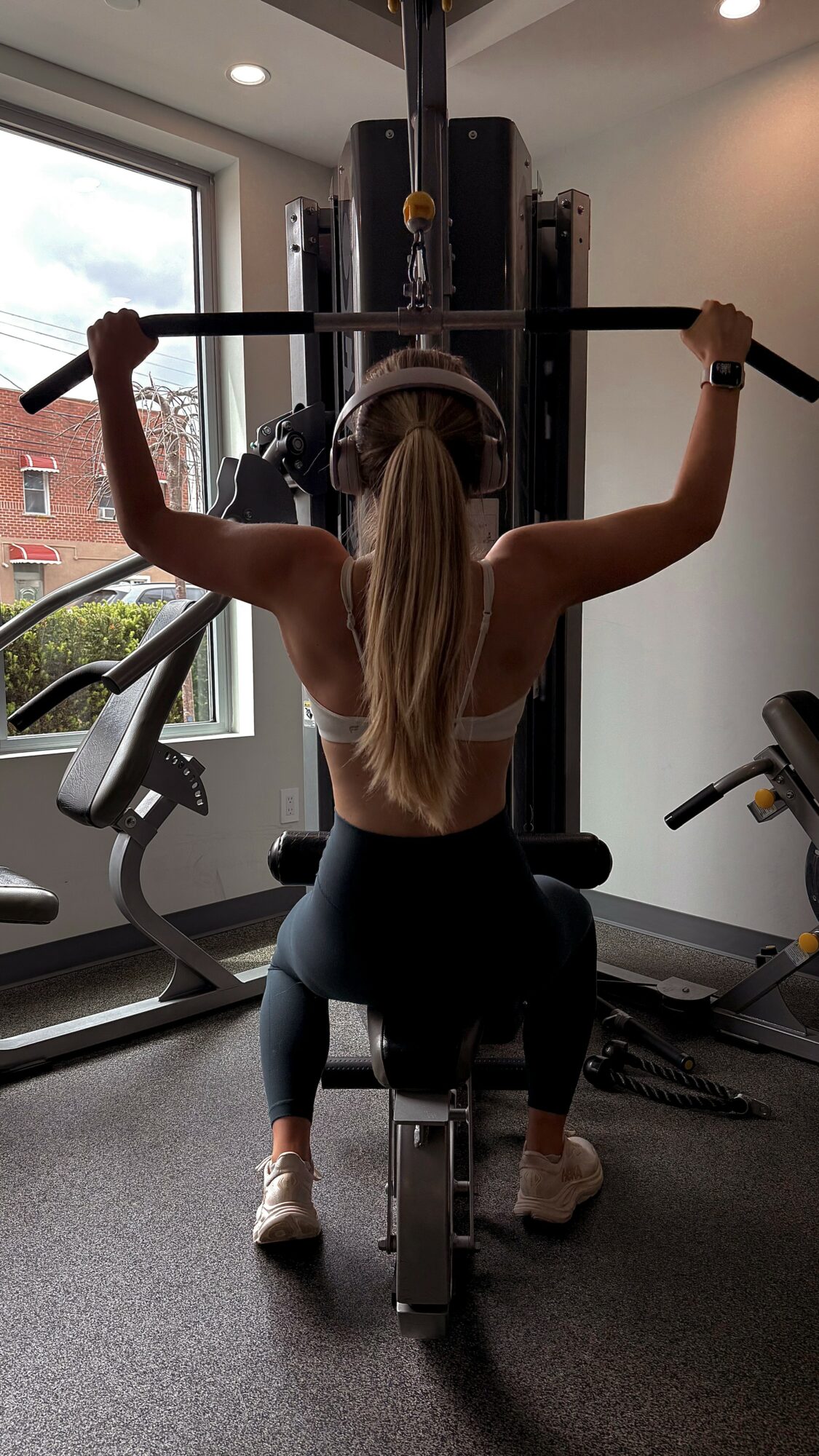 Woman lifting a barbell in a gym, facing away, with gym equipment and window in background.
