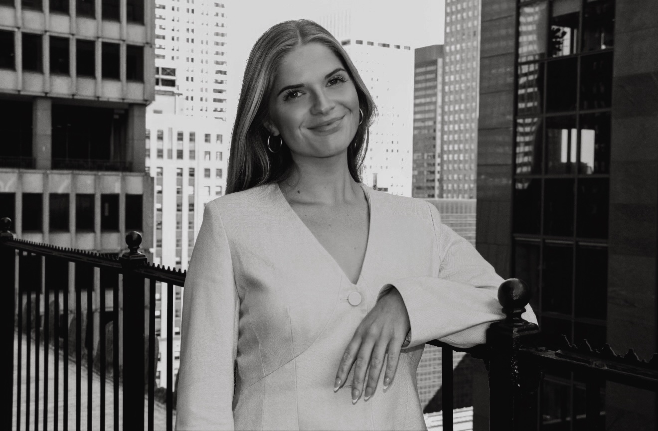 Woman with long hair smiling, standing outdoors in an urban setting with tall buildings, railing, and cityscape behind her.