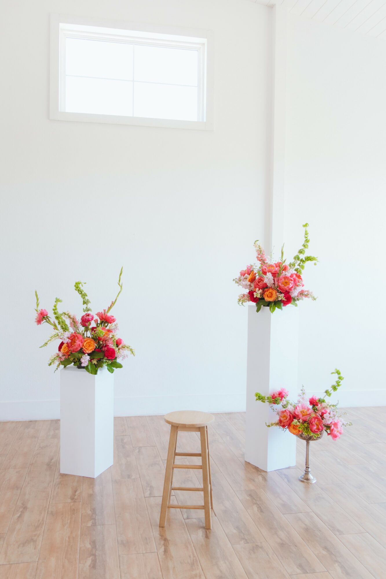 Three flower arrangements in white vases on wooden pedestals and floor, with a window on a white wall in the background.