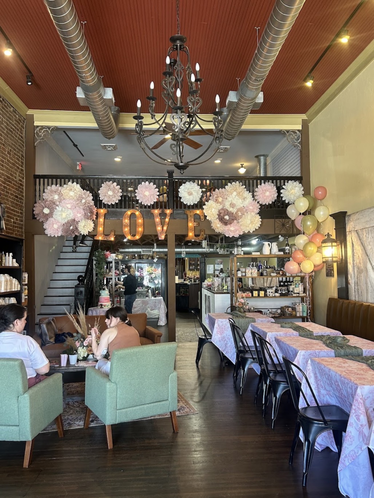 Interior of a cafe with a chandelier, love sign, balloons, tables, and chairs, with people sitting and chatting.