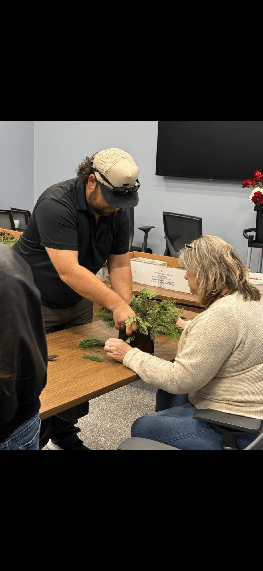 Person handing a plant to a seated woman in a room with a large screen and flowers.