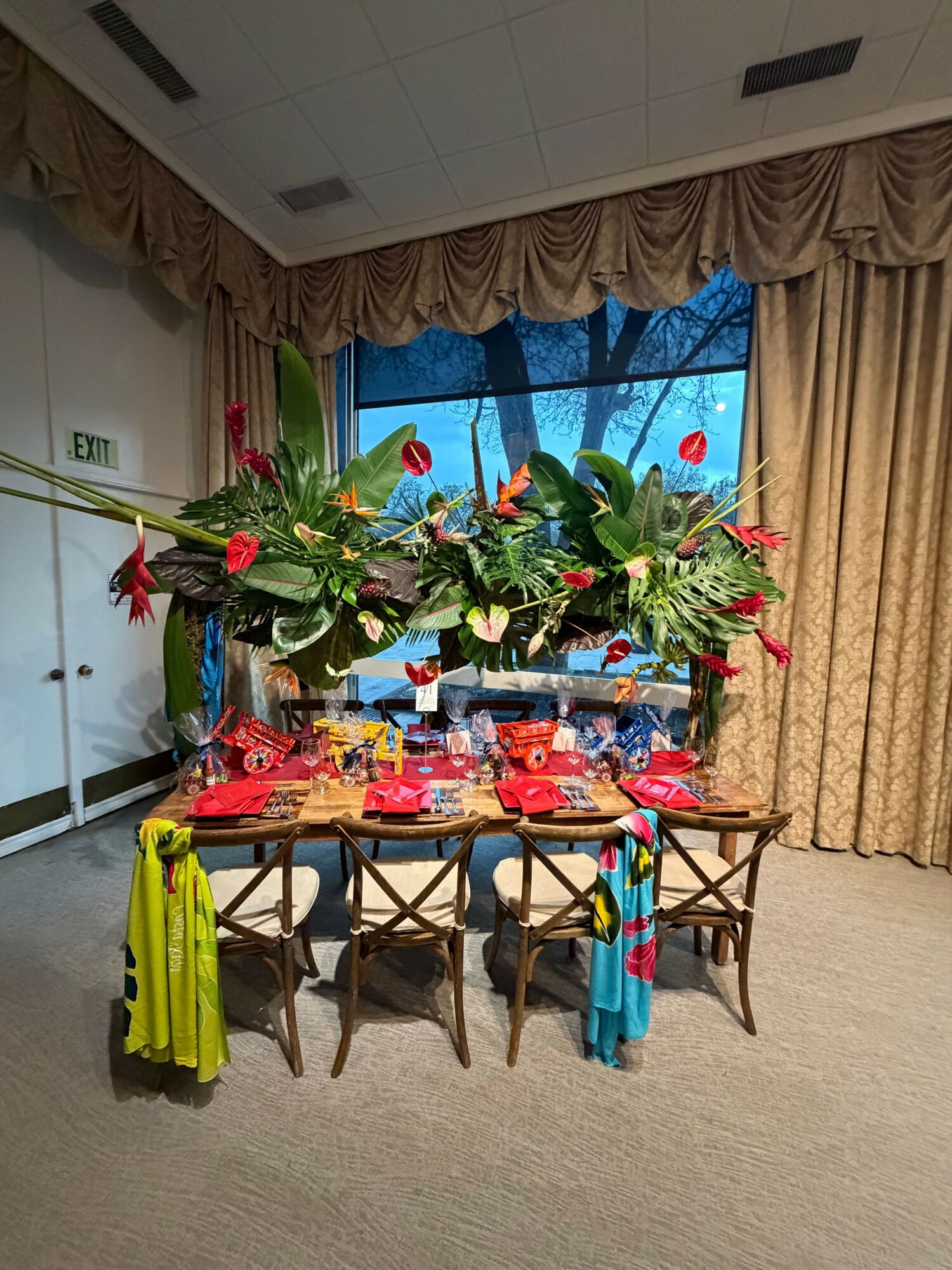 Dining table with colorful decorations and a large floral centerpiece, surrounded by chairs, in a room with curtains and a window.