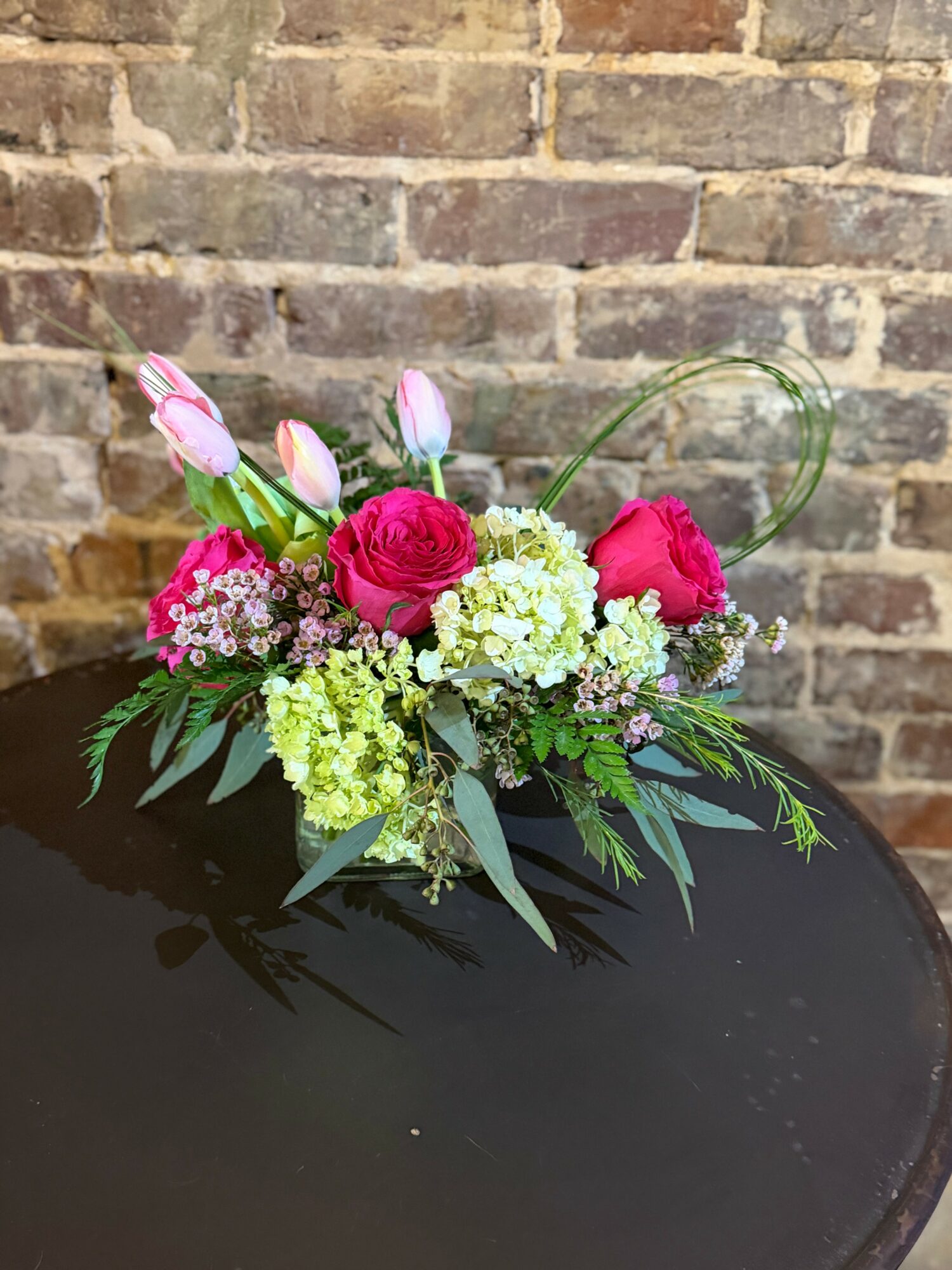Colorful flower arrangement with pink, white, and green flowers on a black surface against a brick wall background.