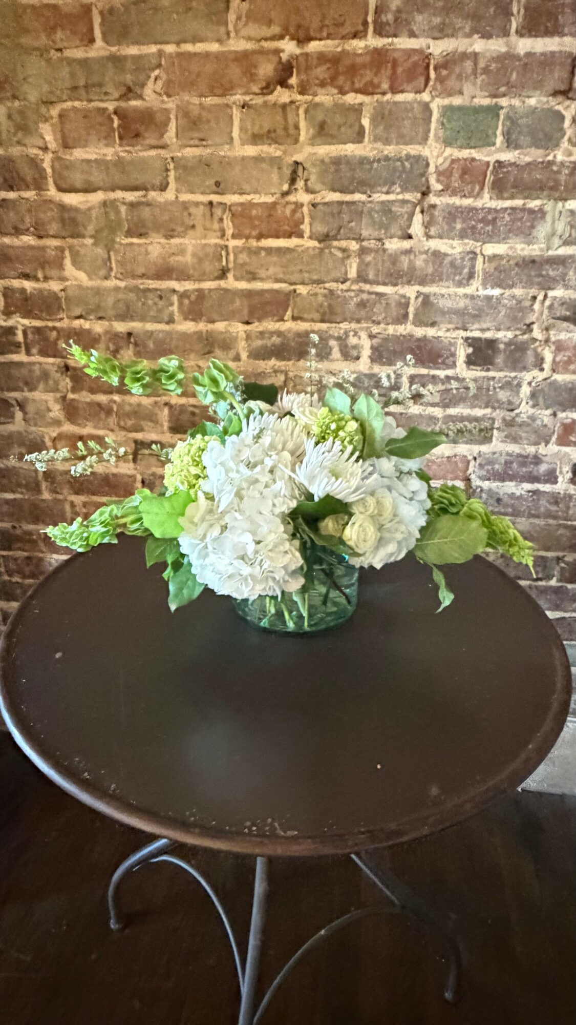 Flower arrangement with white and green flowers on a dark round table against a brick wall background.