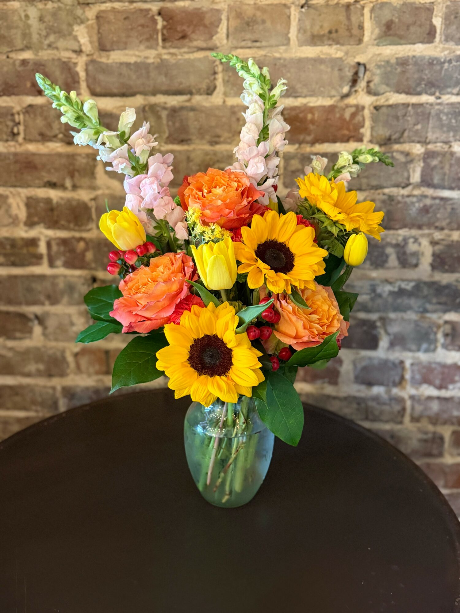 Colorful flower bouquet with sunflowers, pink, orange, and yellow flowers in a green glass vase against a brick wall.