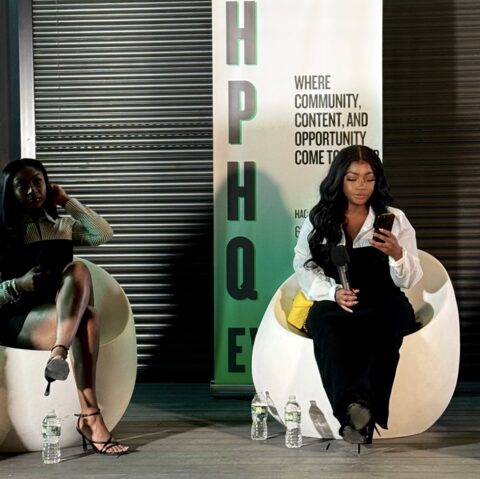 Two women sit on modern chairs, one looking at her phone, with a vertical sign reading 'HQE' and text about community and opportunity.