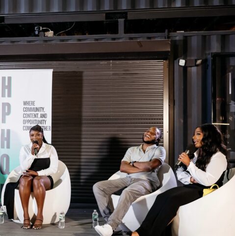 Three people seated on white chairs, engaging in a discussion, with a black background and a partially visible sign.