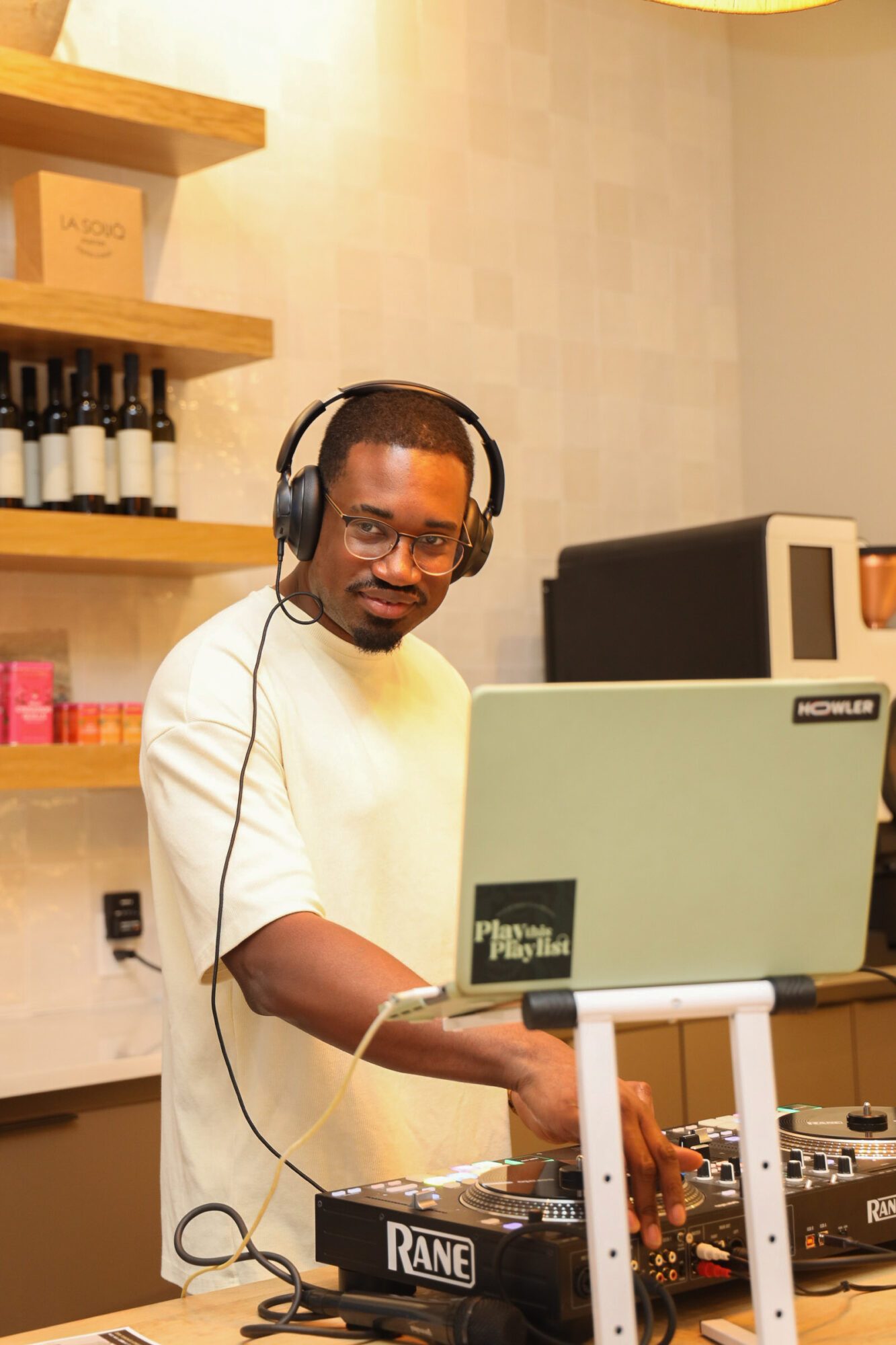 Person wearing headphones working with DJ equipment in a room with shelves of bottles and a computer.