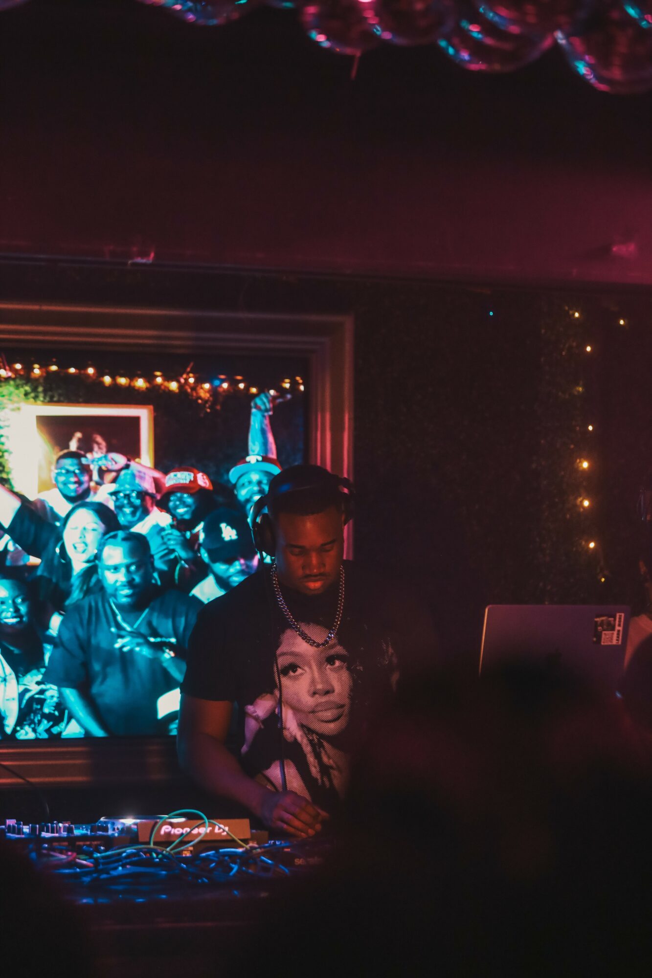 DJ wearing a black T-shirt with a woman's face, standing in front of a screen showing a group of people, in a dimly lit room.