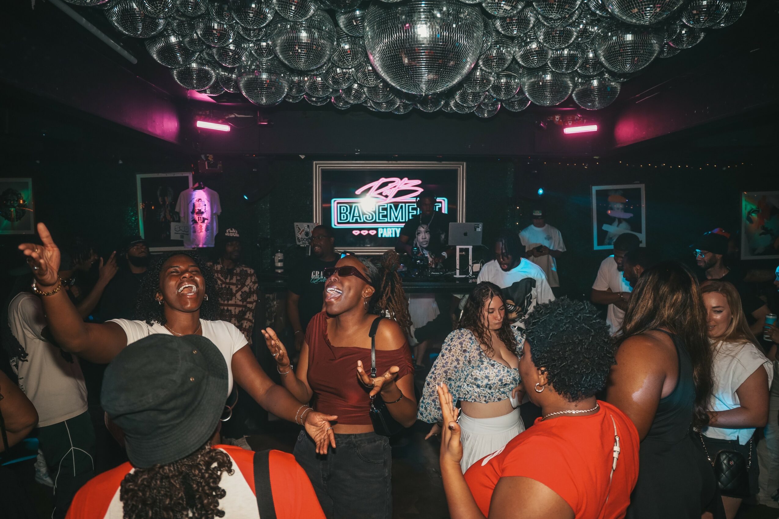 People dancing and socializing in a nightclub with a DJ and colorful lighting, ceiling decorations, and a large screen in the background.