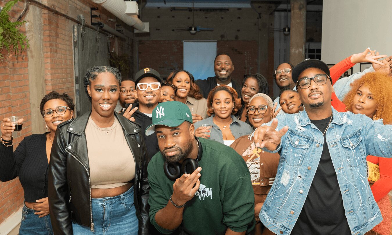 Group of diverse people smiling and posing indoors, some making gestures, with brick and wooden walls in background.