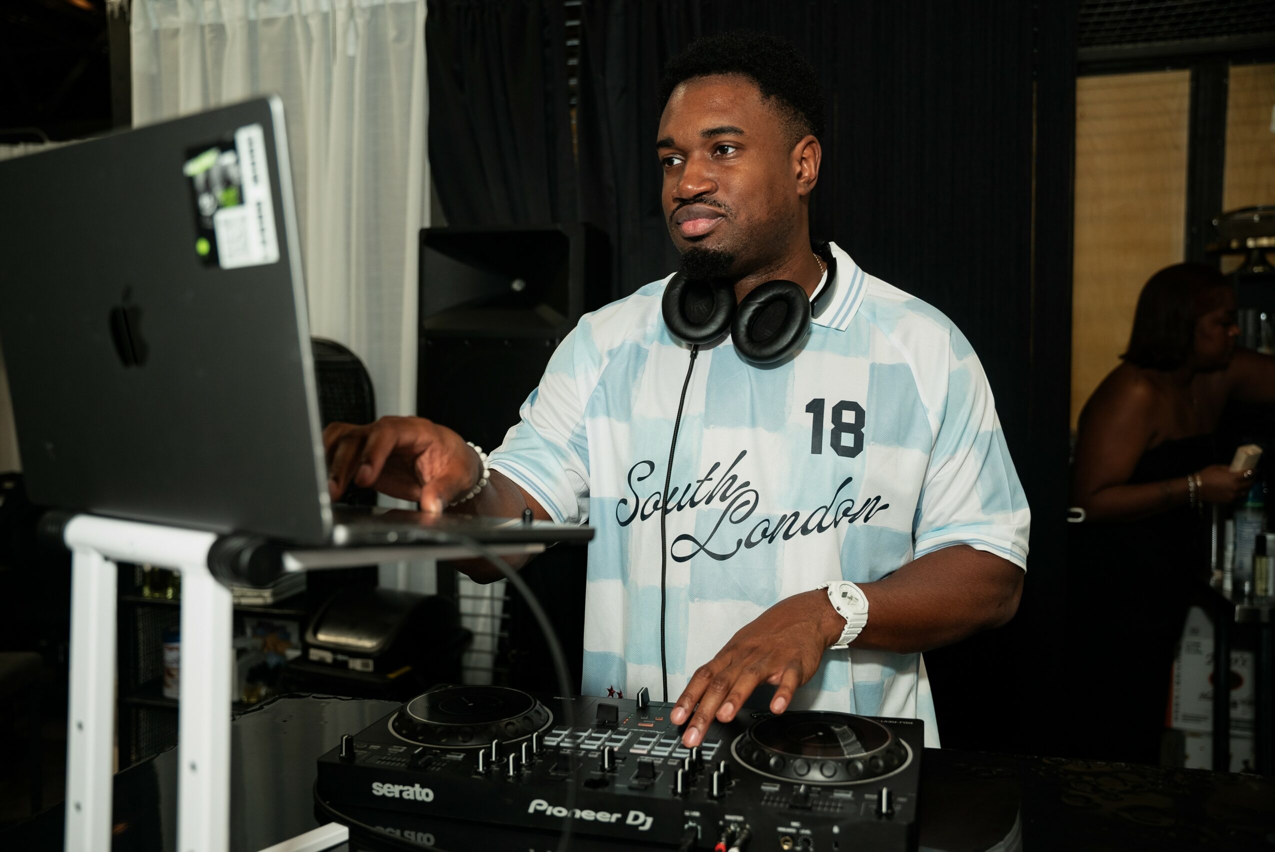 DJ wearing headphones and a striped shirt, working at a DJ setup with a laptop and equipment, in a dimly lit room.