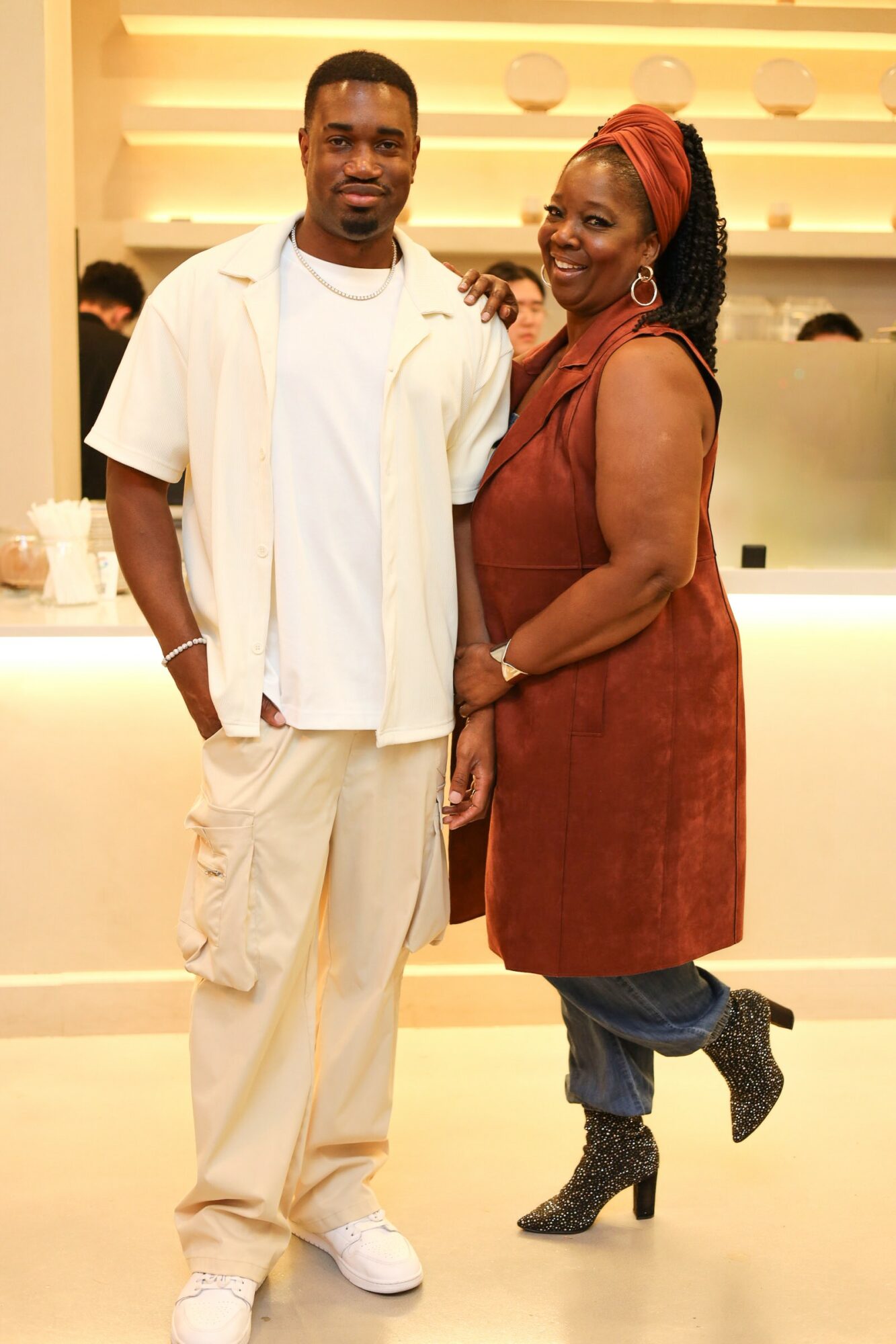 A man and woman stand together indoors, smiling, with a brightly lit background and a counter behind them.