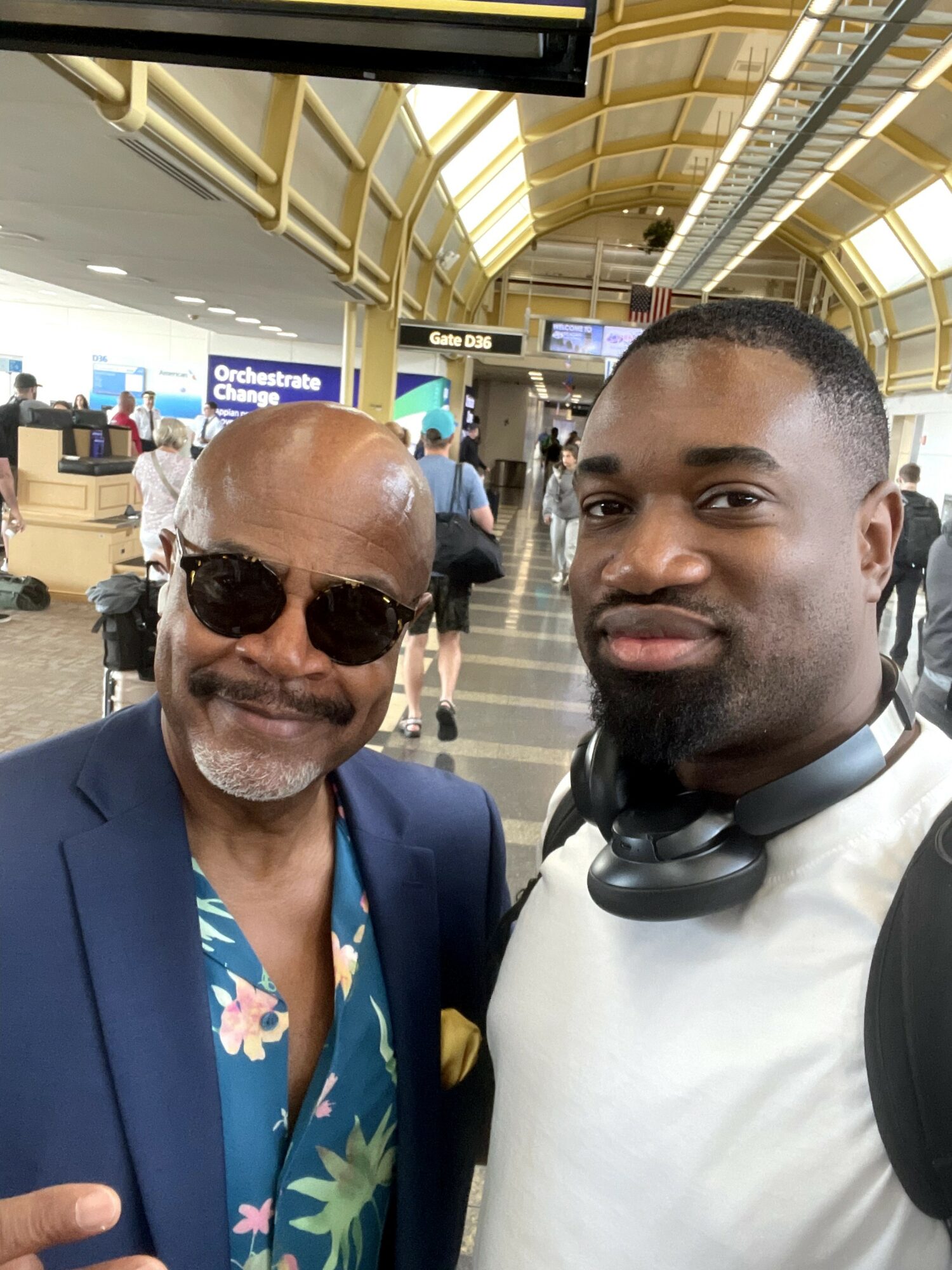 Two men taking a selfie in an airport terminal with a curved ceiling and people in the background.