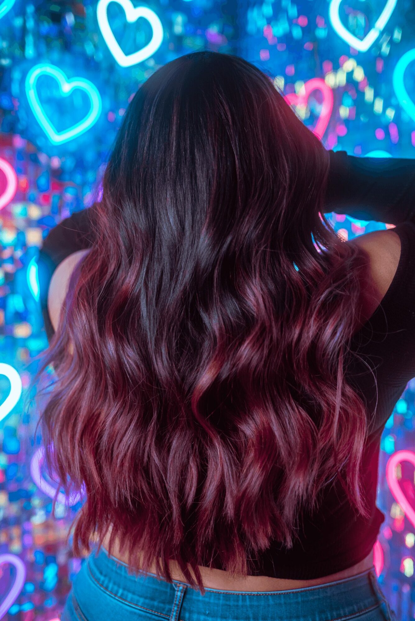 Back of a person with long, wavy hair dyed in shades of dark purple and pink, against a colorful heart-themed background.