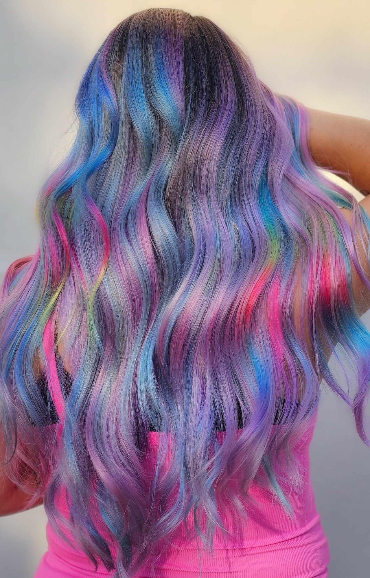 Person with long, wavy hair dyed in vibrant shades of purple, blue, pink, and silver, flowing down their back.
