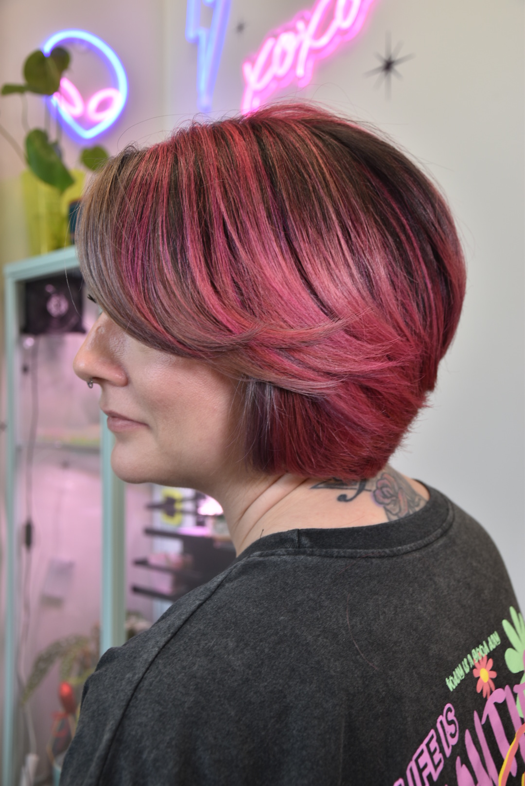 Side view of a person with short, pinkish-red hair in a salon, neon signs in the background.