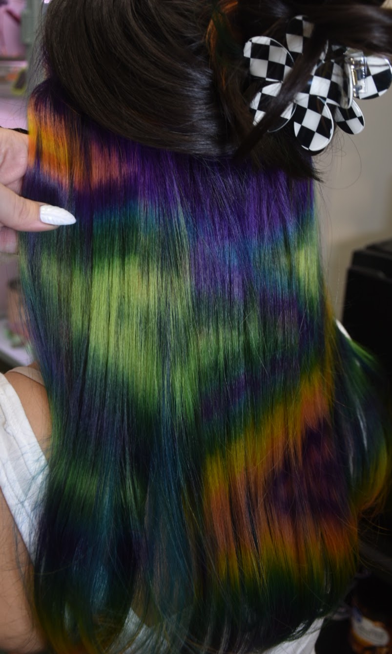 Close-up of long, straight, multicolored hair with green, purple, blue, and orange shades, held by a hand with white nails, with a checkered bow in the background.