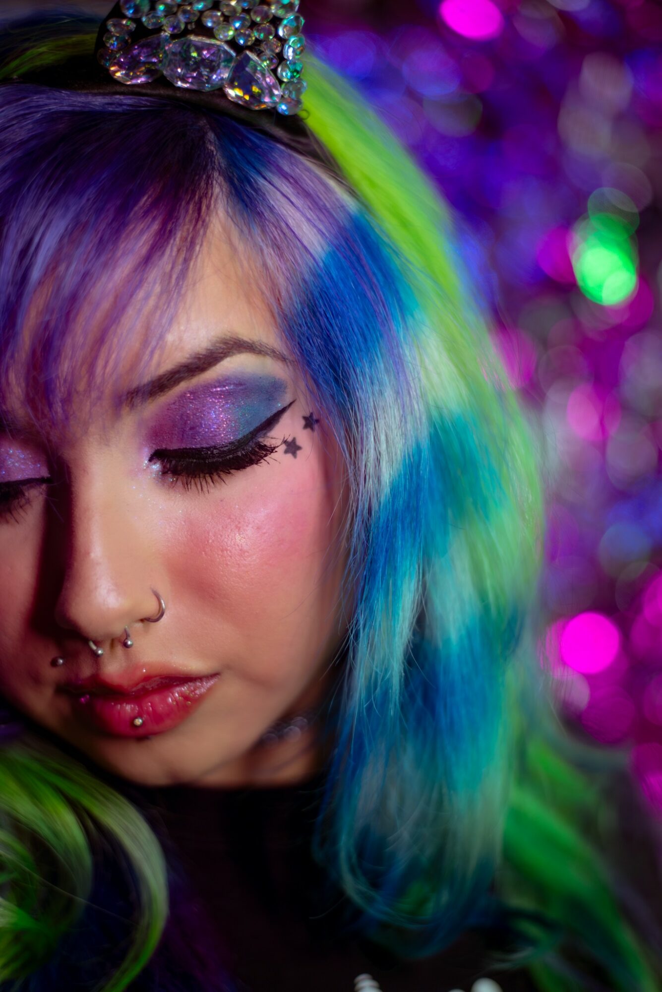 Close-up of a person with colorful hair, makeup, and a small crown, with a blurred colorful background.