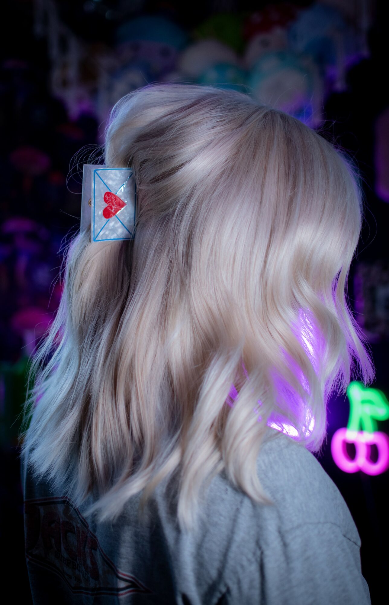Back of a person's head with wavy blonde hair, hair clip with a red heart, colorful background, and neon lights.