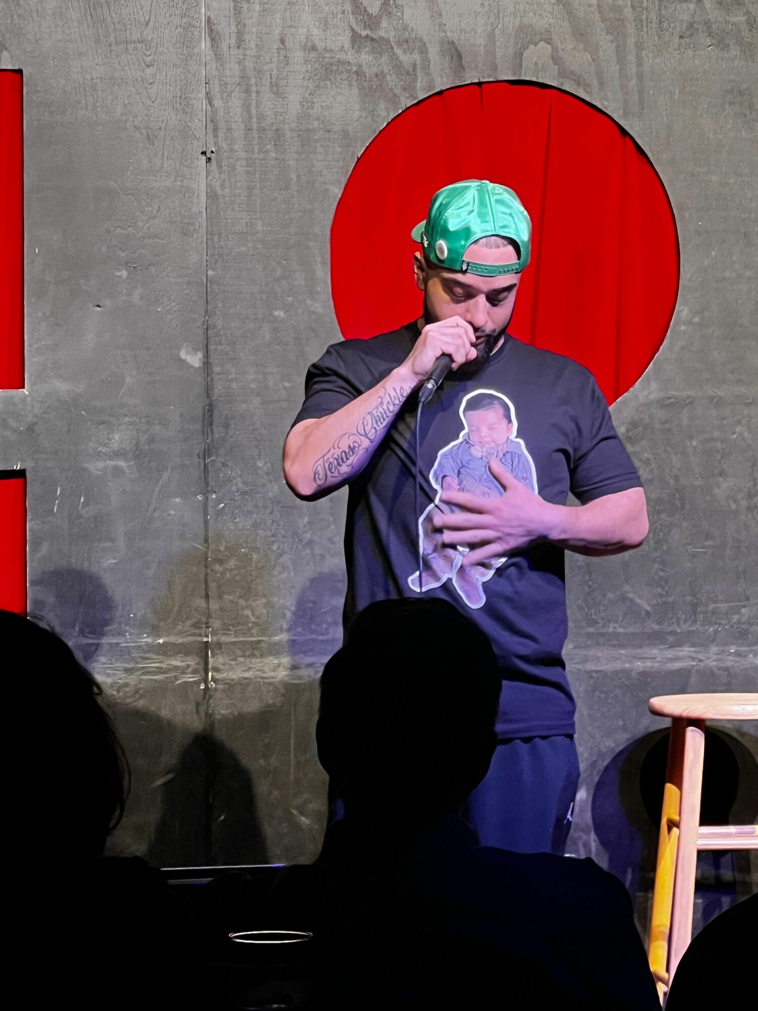 Person wearing a green cap and glasses, talking on a phone, with a red umbrella behind them, against a gray wall.