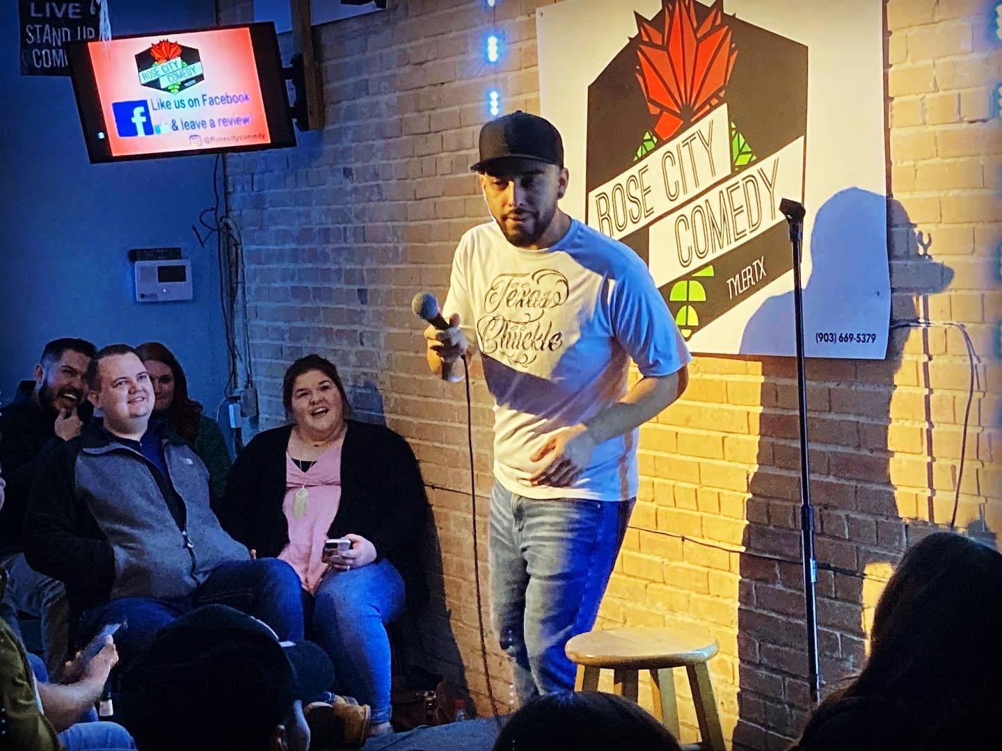 Stand-up comedian holding a microphone on stage with an audience watching, brick wall background, Rose City Comedy sign, and a TV screen.