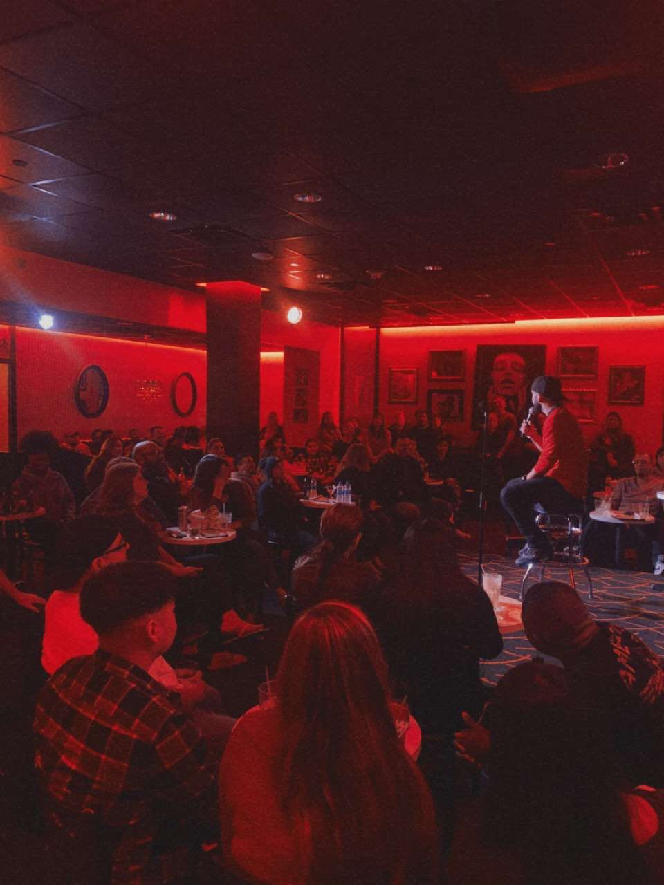 People seated at tables in a dimly lit room with red lighting, some standing and talking, framed by walls with pictures.
