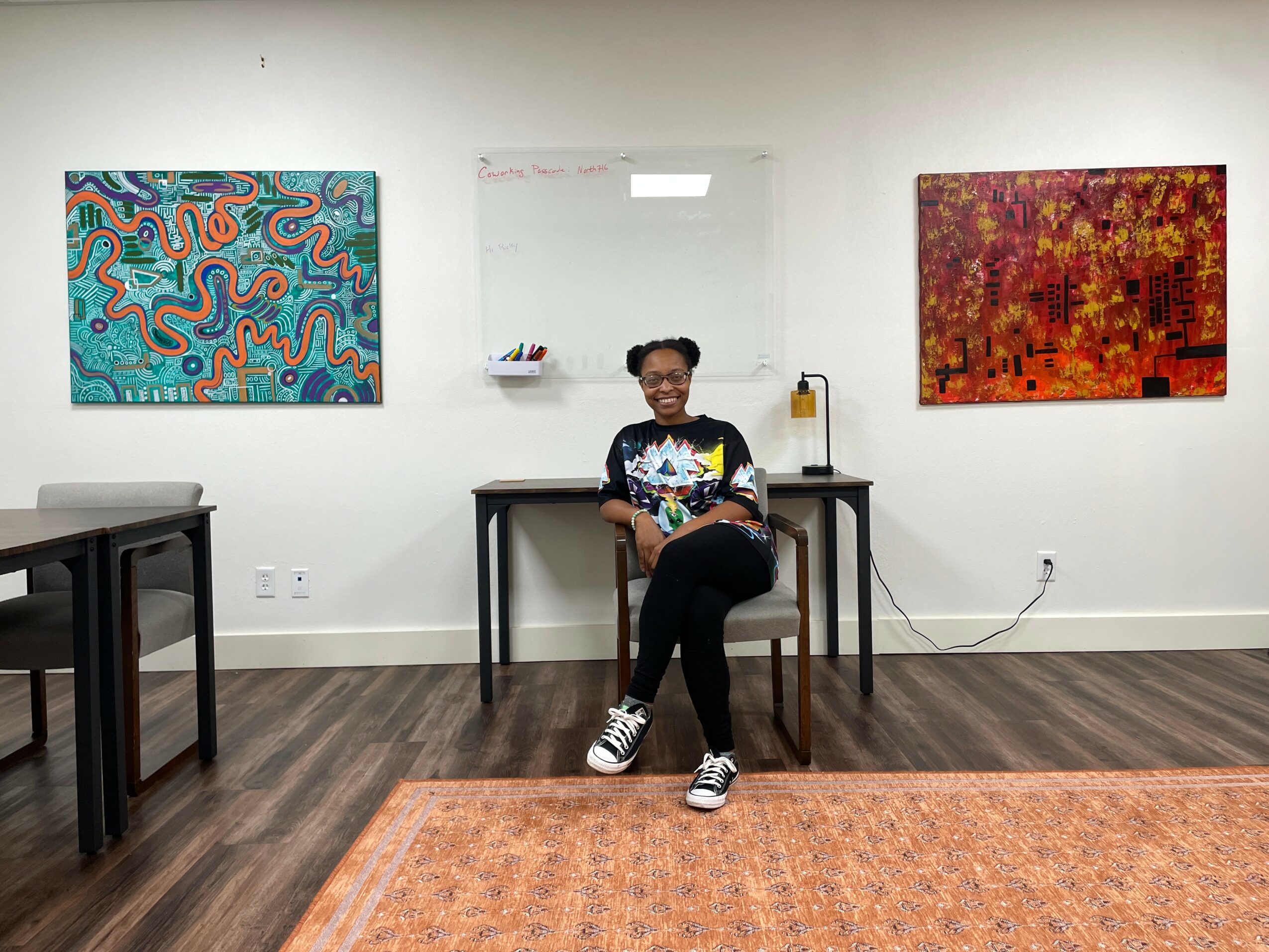 Person sitting on a chair in an art gallery with colorful paintings on white walls, wooden floor, and a whiteboard behind.