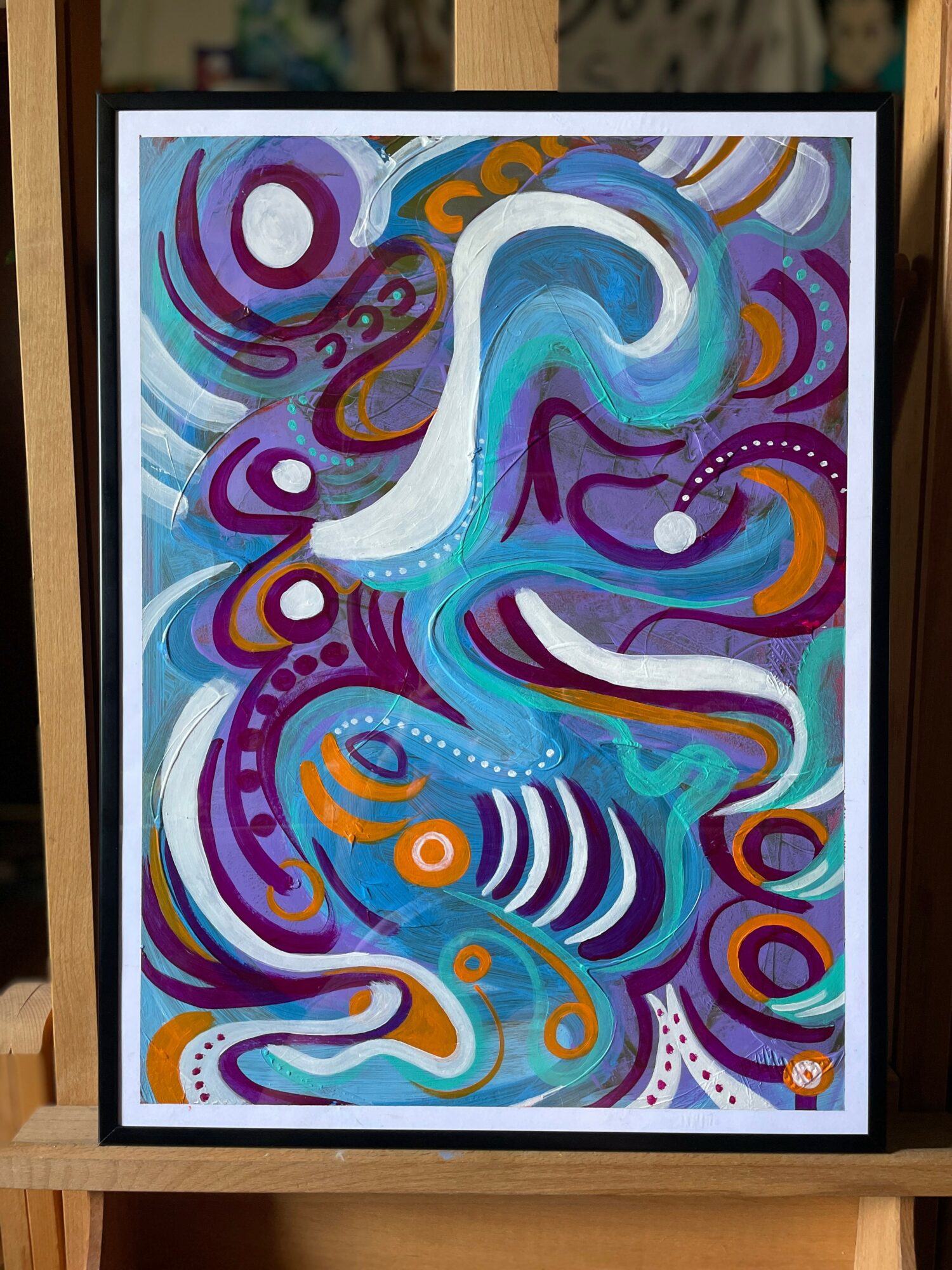 Abstract painting with swirling shapes and vibrant colors including blue, purple, orange, and white, framed and displayed on a wooden surface.