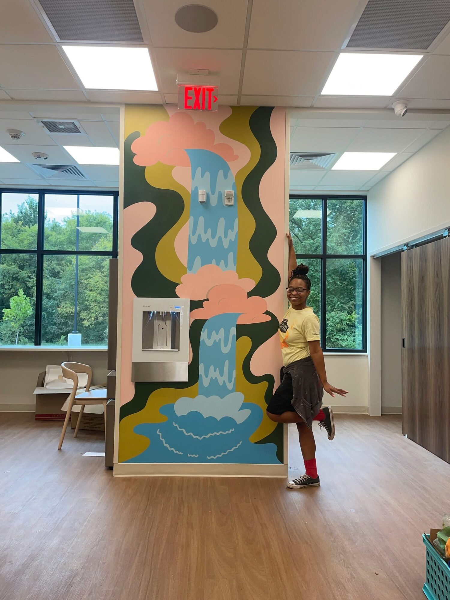 Person standing next to a colorful wall mural of a waterfall with trees and clouds, in a room with large windows.