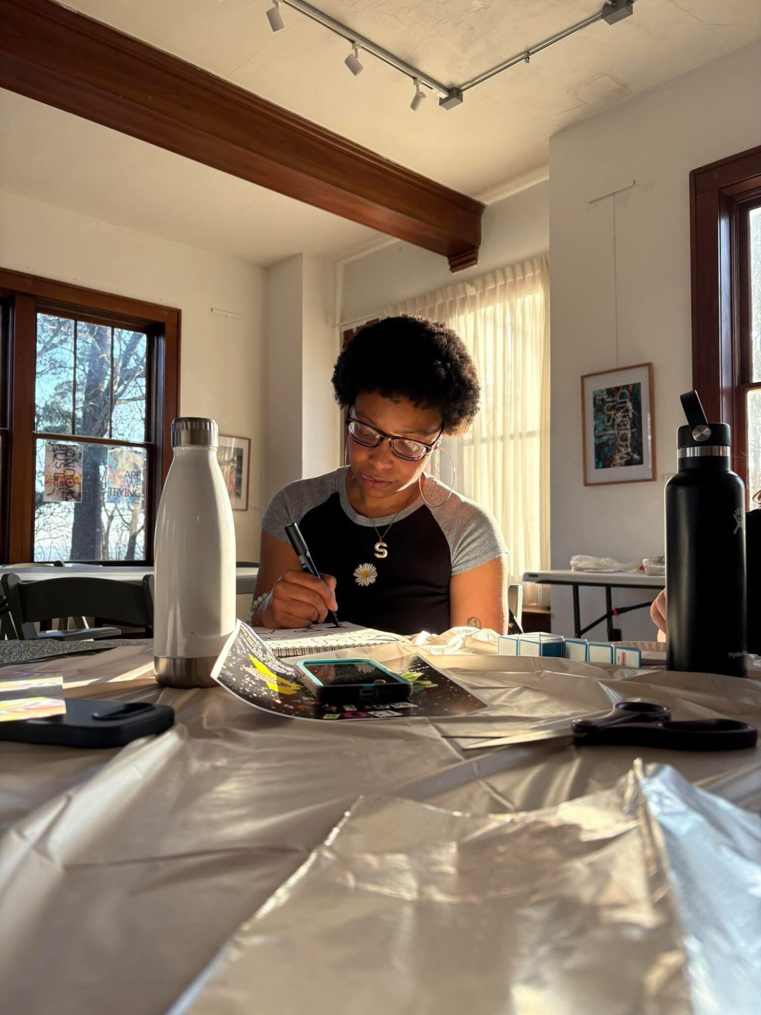 Young person with curly hair and glasses writing at a table with water bottles and papers, sunlight streaming through windows.