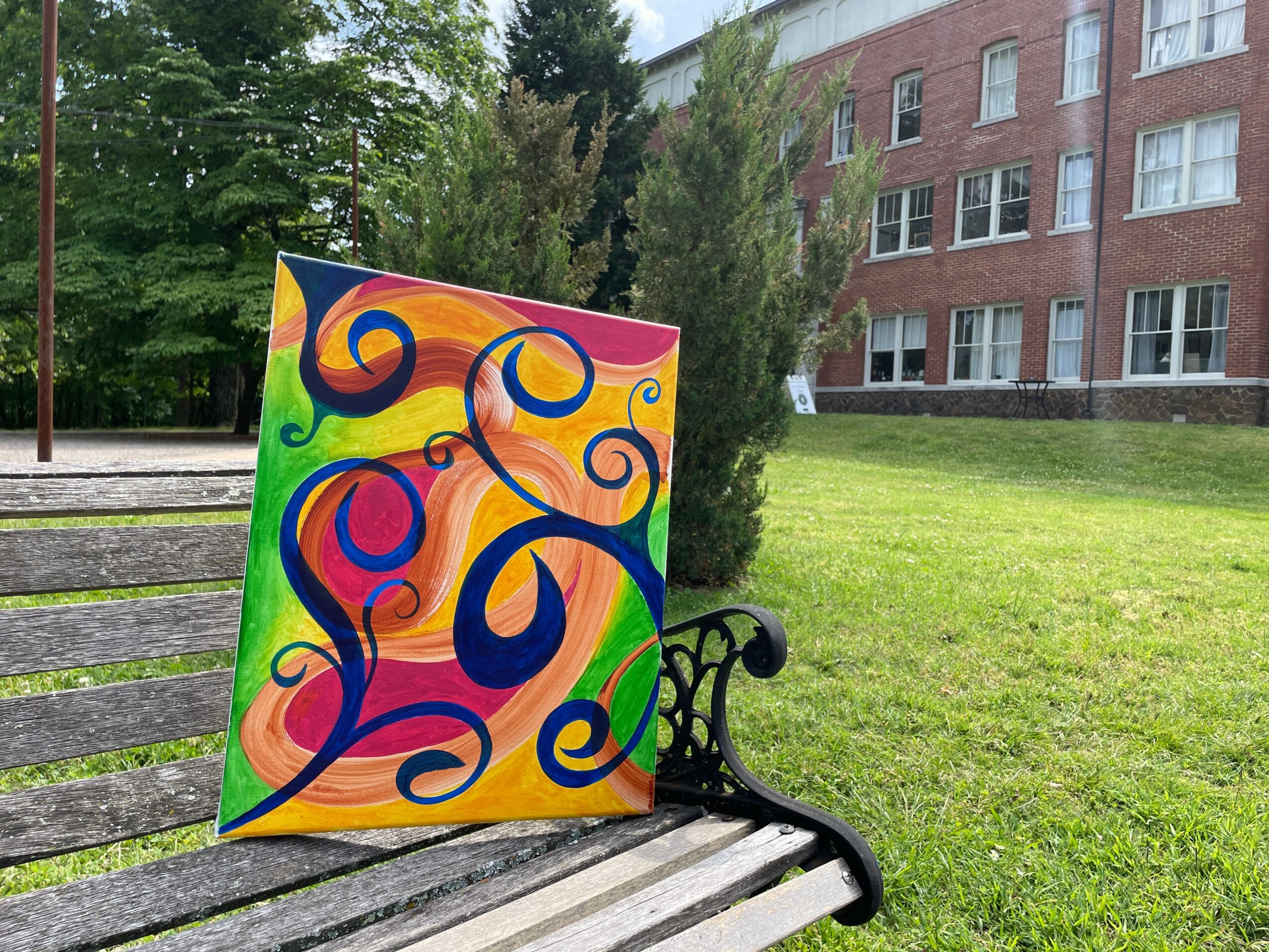 Colorful abstract painting with swirls and shapes leaning against a park bench, with trees and a building in the background.