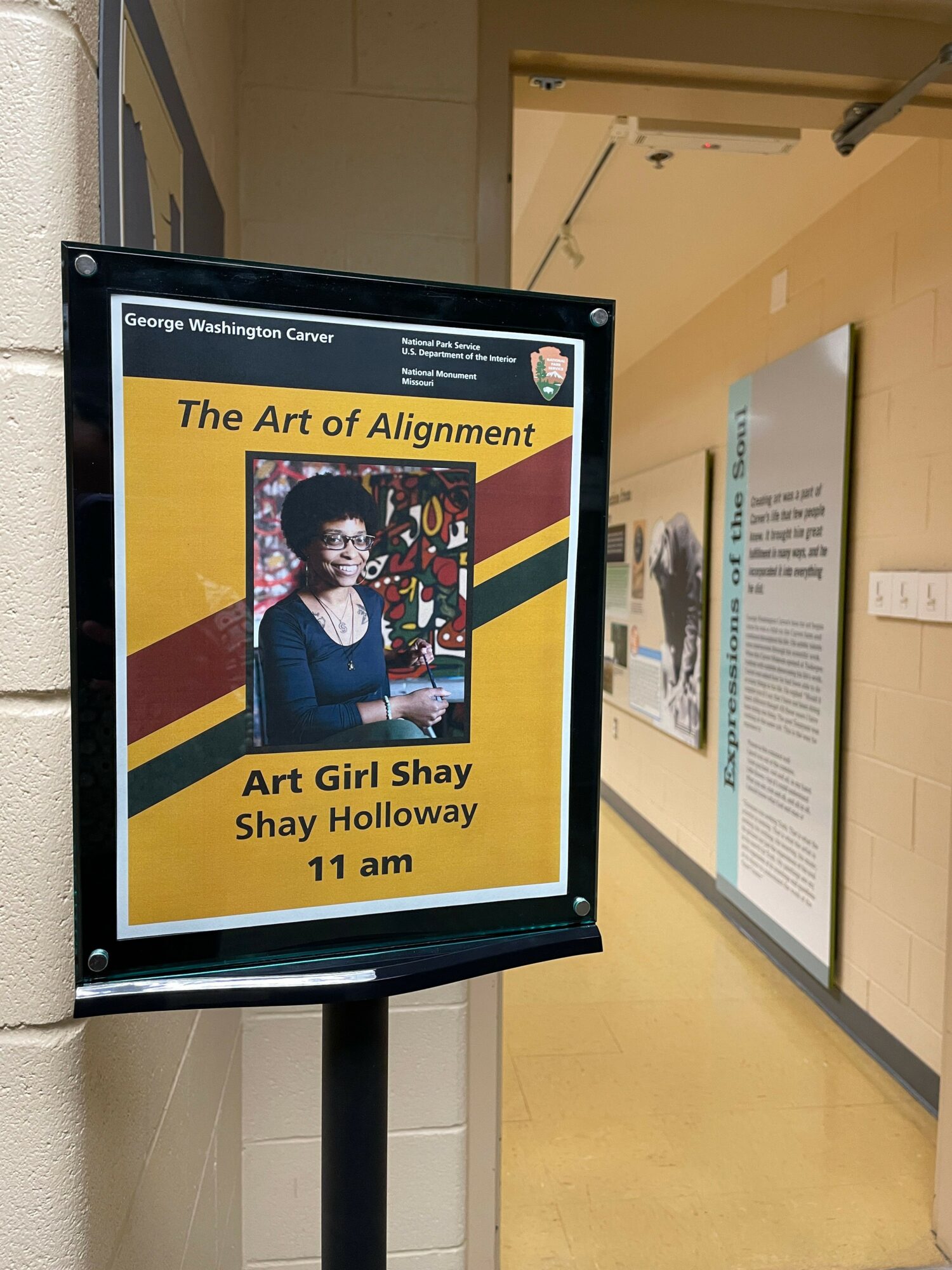 Digital display showing event details for Art Girl Shay and Shay Holloway at 11 am, with yellow and maroon background.