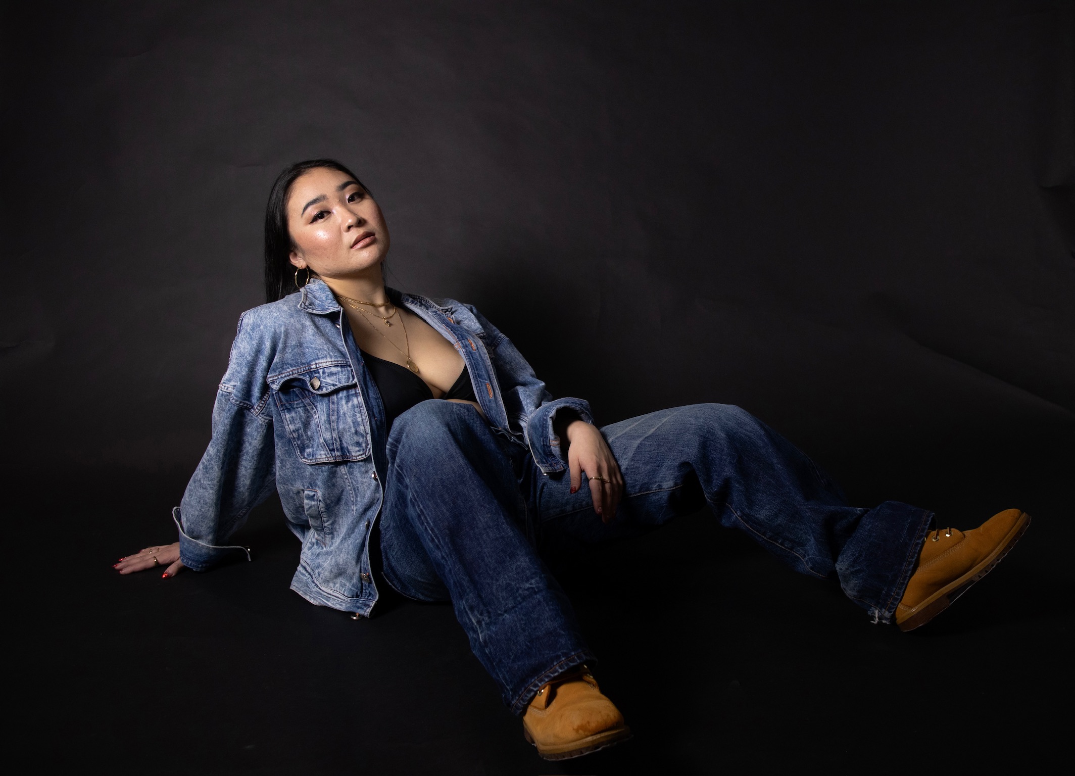 Woman in denim jacket and jeans sitting on black background, looking at camera, relaxed pose, yellow shoes, dark hair.