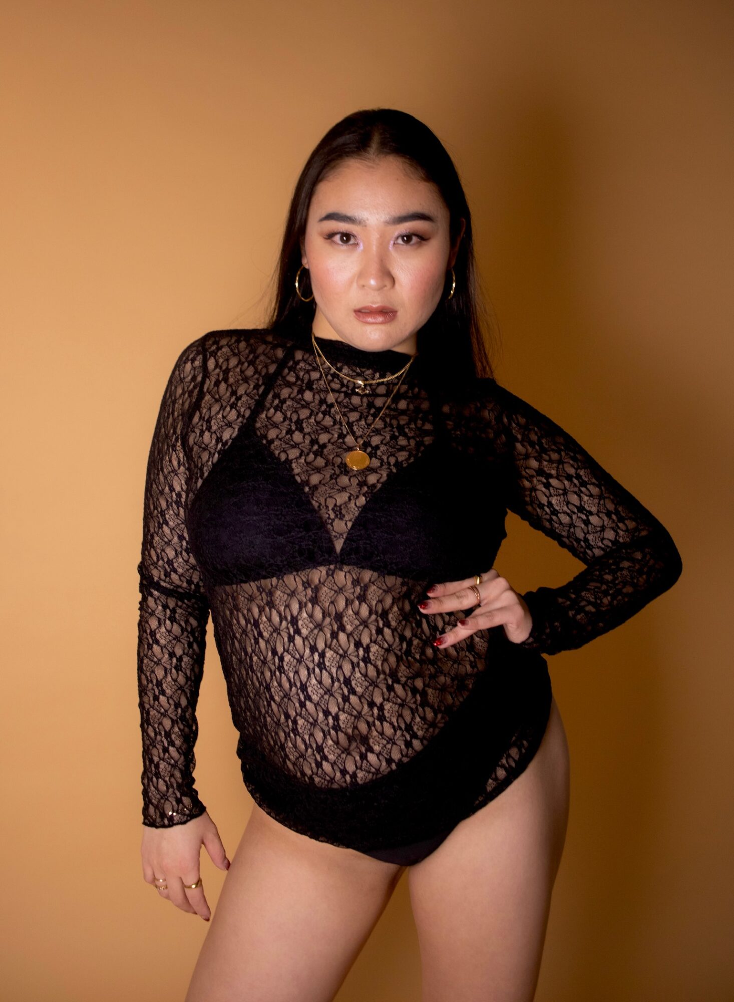 Woman in black lace outfit with a yellow background, posing with hand on hip, looking at camera.