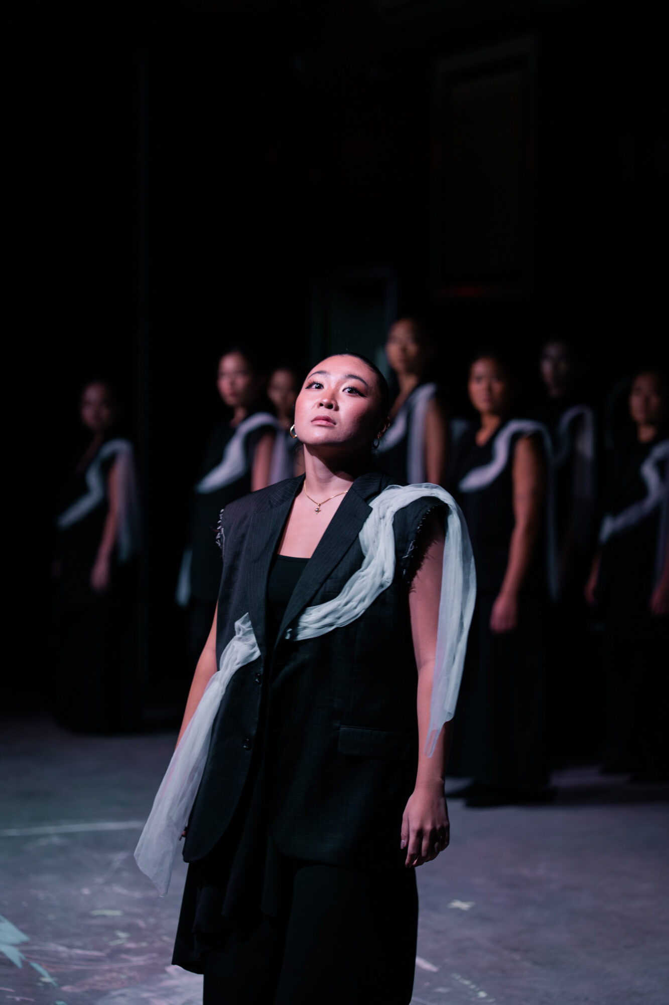 A woman in a black outfit stands in front of a group of women dressed similarly, in a dark setting.