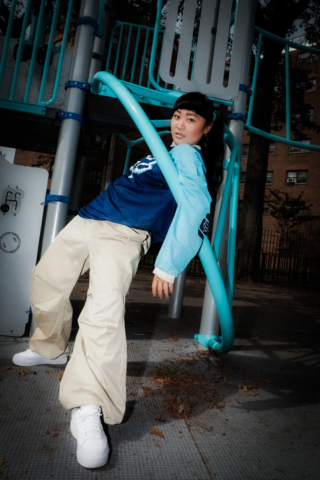 Young person leaning on playground equipment at night, wearing a blue and light blue jacket, beige pants, and white shoes.