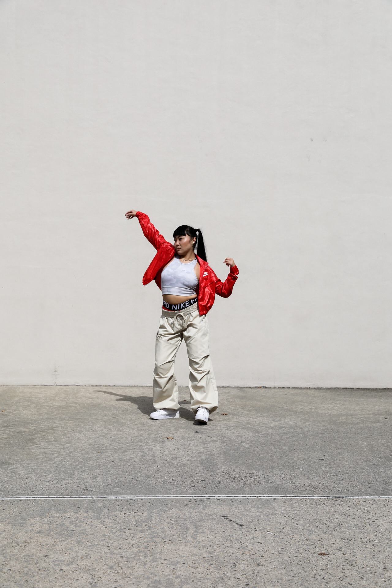 Person standing against a plain wall, wearing a red jacket, crop top, and light-colored pants, with arms raised.