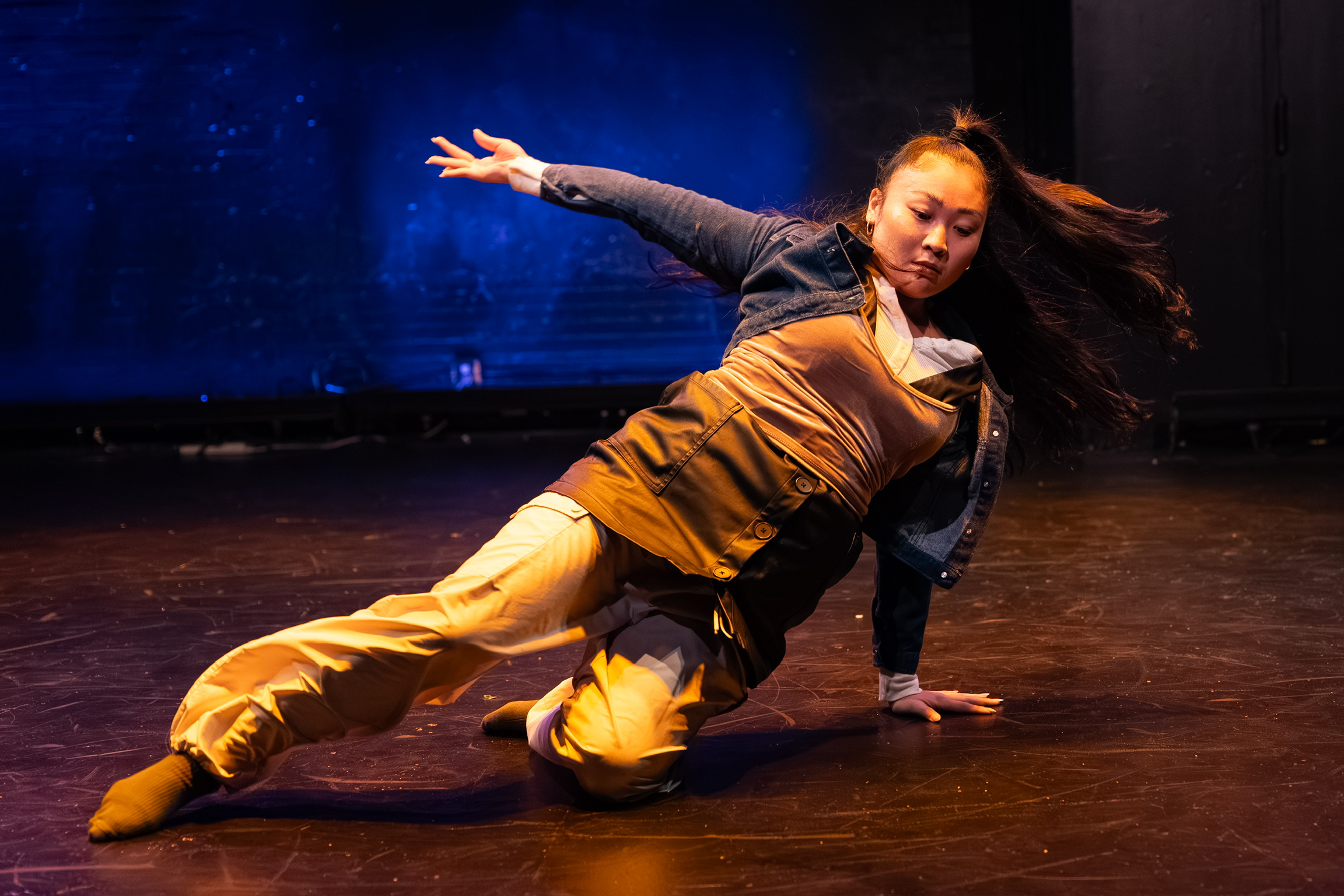 A woman performs a dance move on stage, leaning to one side with one hand on the floor and hair flowing.