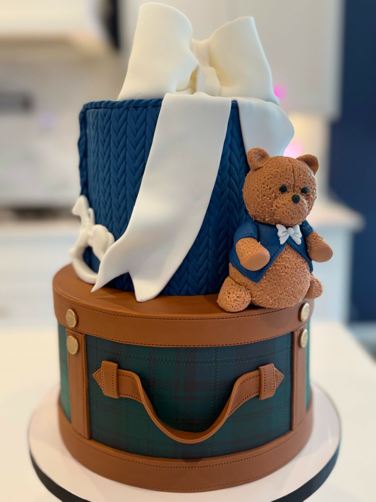 Cake decorated as a suitcase with a teddy bear and tissue box on top, with a brown handle design, neutral background.
