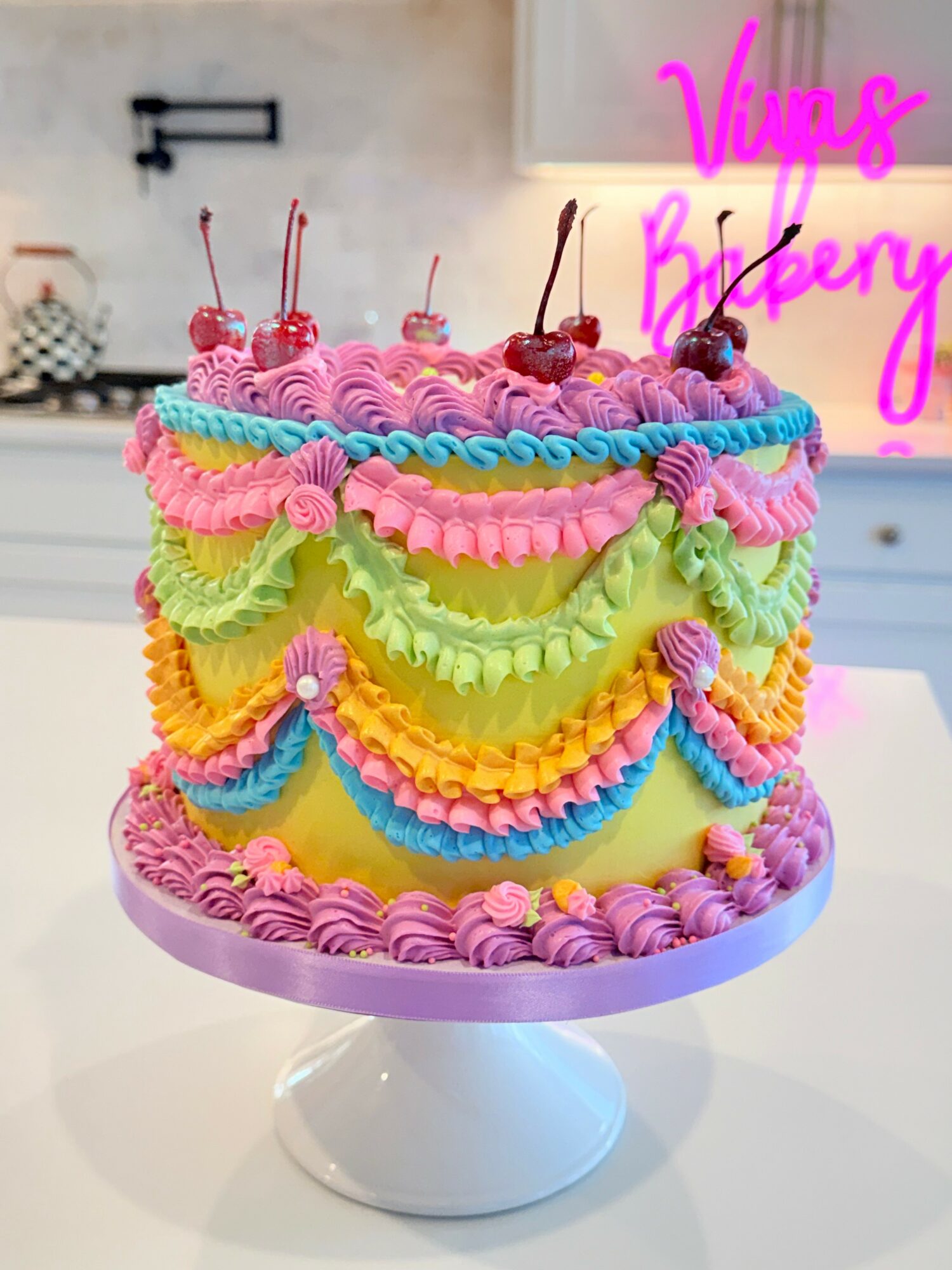 Colorful layered cake with pink, green, yellow, and blue frosting, topped with cherries, on a white cake stand.