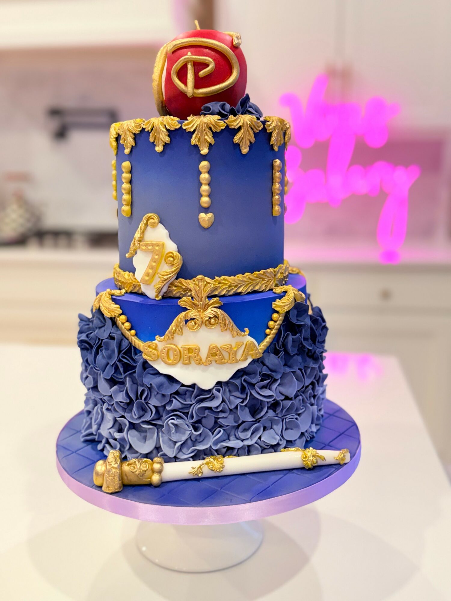 Two-tiered cake with blue and gold decorations, topped with a red and gold emblem, and a gold chain with a scroll.