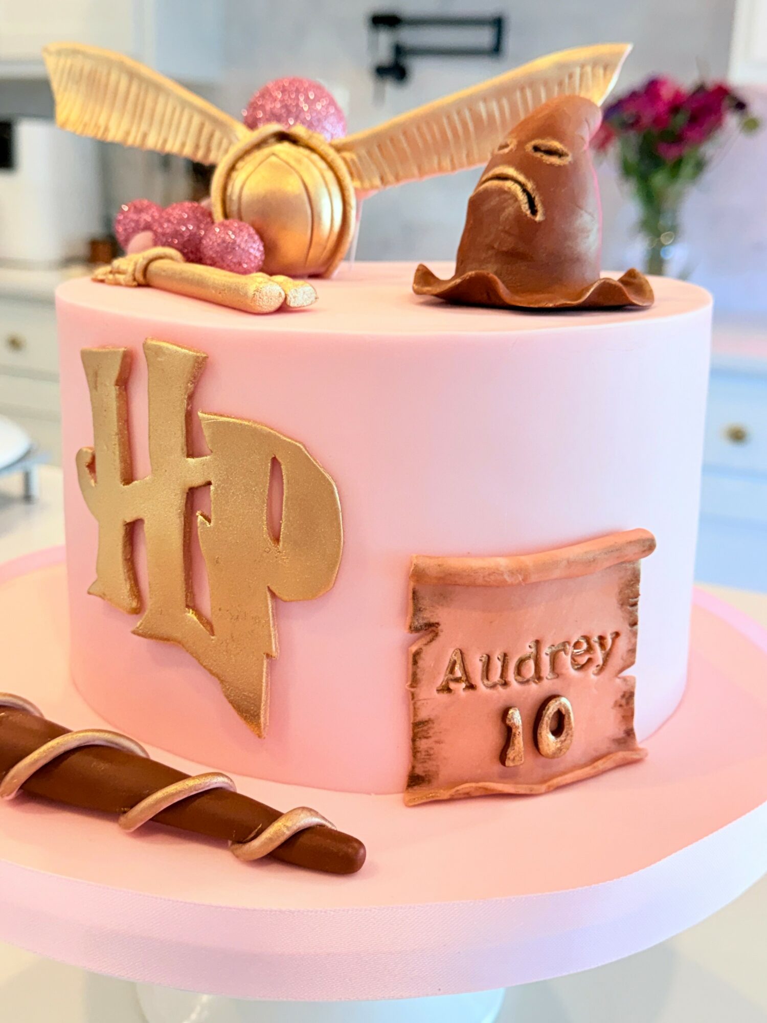 Pink birthday cake with Harry Potter-themed decorations, including a golden Harry Potter logo, a chocolate frog, and a scroll with 'Audrey 10' written.