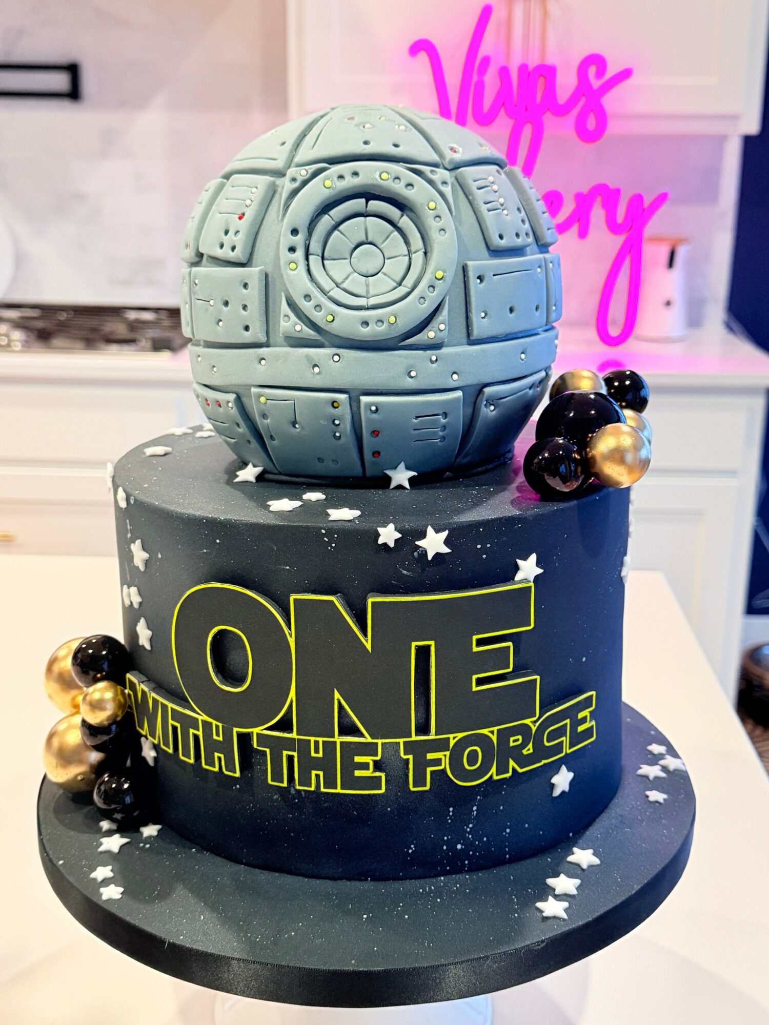 A two-tiered cake with a space theme, featuring a large spherical spaceship on top and star decorations, on a black cake stand.