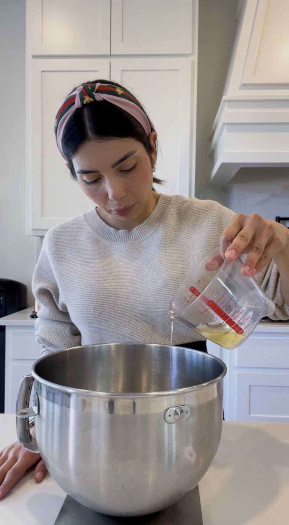 Woman pouring ingredients into a large metal mixing bowl in a kitchen, wearing a headband and light sweater.