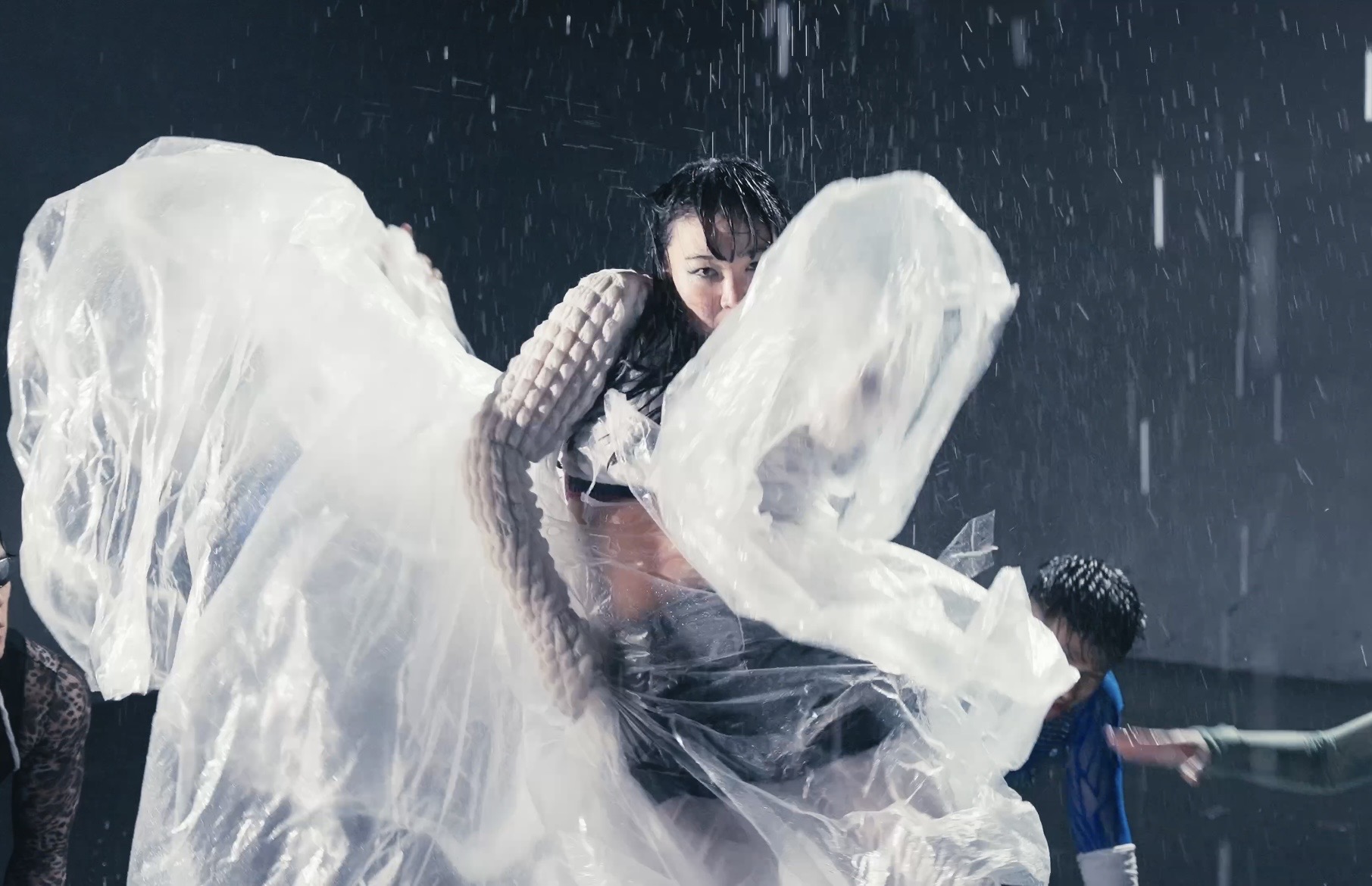 Person in rain with large plastic sheet, others nearby, dark background, rain falling heavily.
