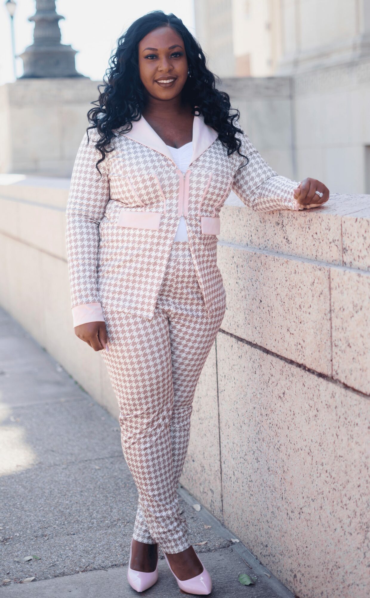 Woman with long curly hair smiling, wearing a checkered suit, standing outdoors near a stone wall.